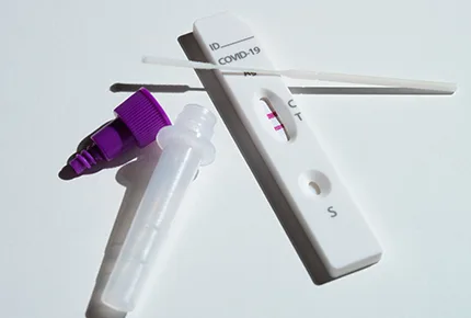 Close-up of a covid-19-test kit showing a positive result, with a test strip, dropper, and swab placed on a white surface.